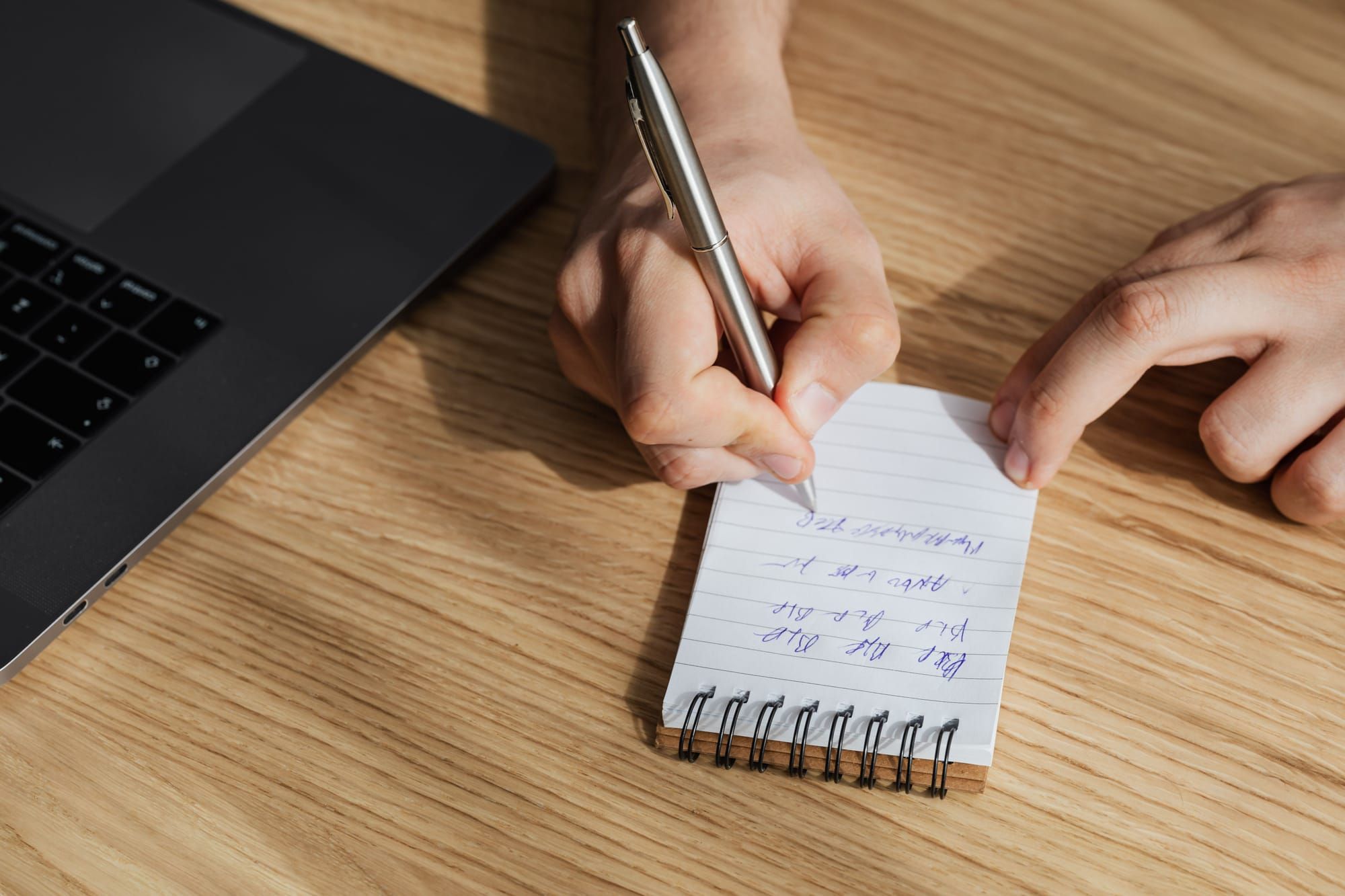 Mano escribiendo con un bolígrafo en una libreta pequeña junto a un portátil, sobre una mesa de madera