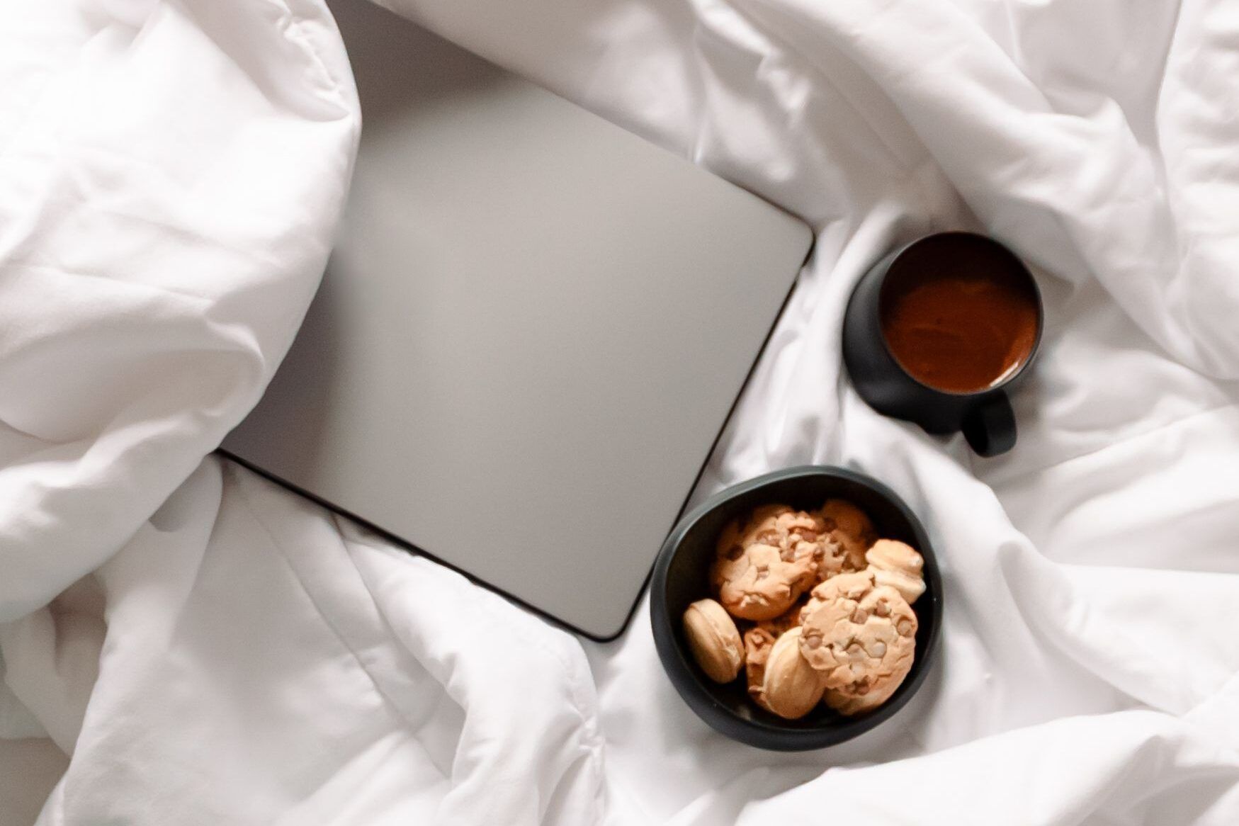 Portátil cerrado sobre una cama junto a un cuenco de galletas y una taza de café, vistos desde arriba