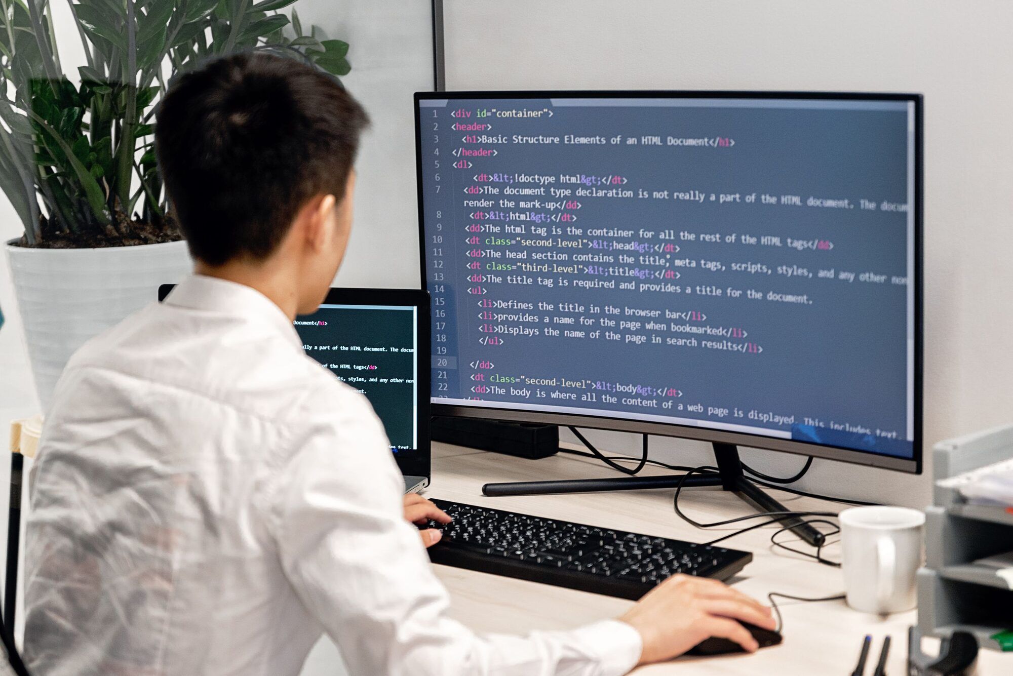Técnico de espaldas trabajando ante un monitor con código, con un portátil y una taza en la mesa