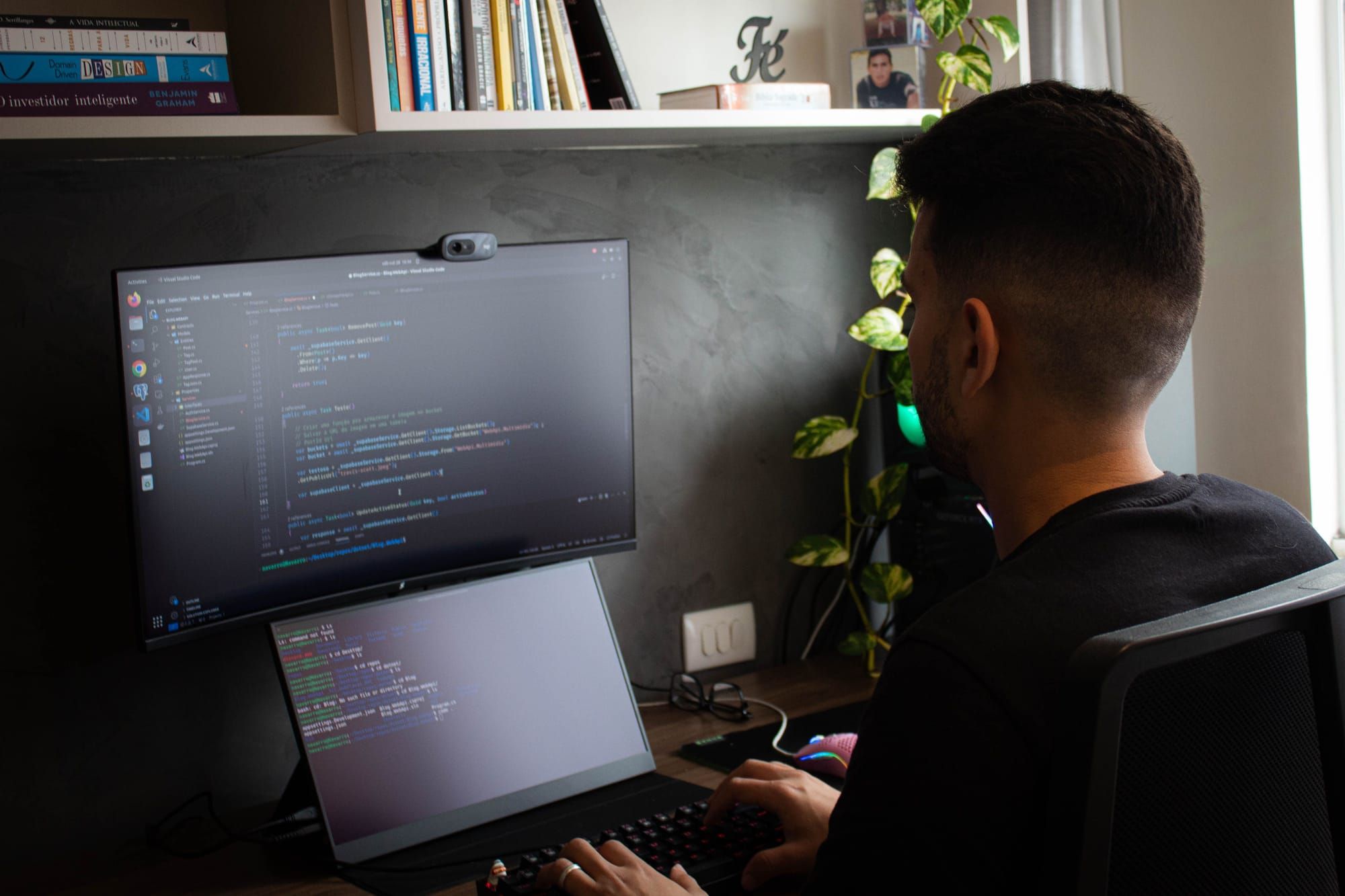 Hombre de espaldas en su mesa con código en un monitor y una terminal en el portátil, junto a una estantería con libros