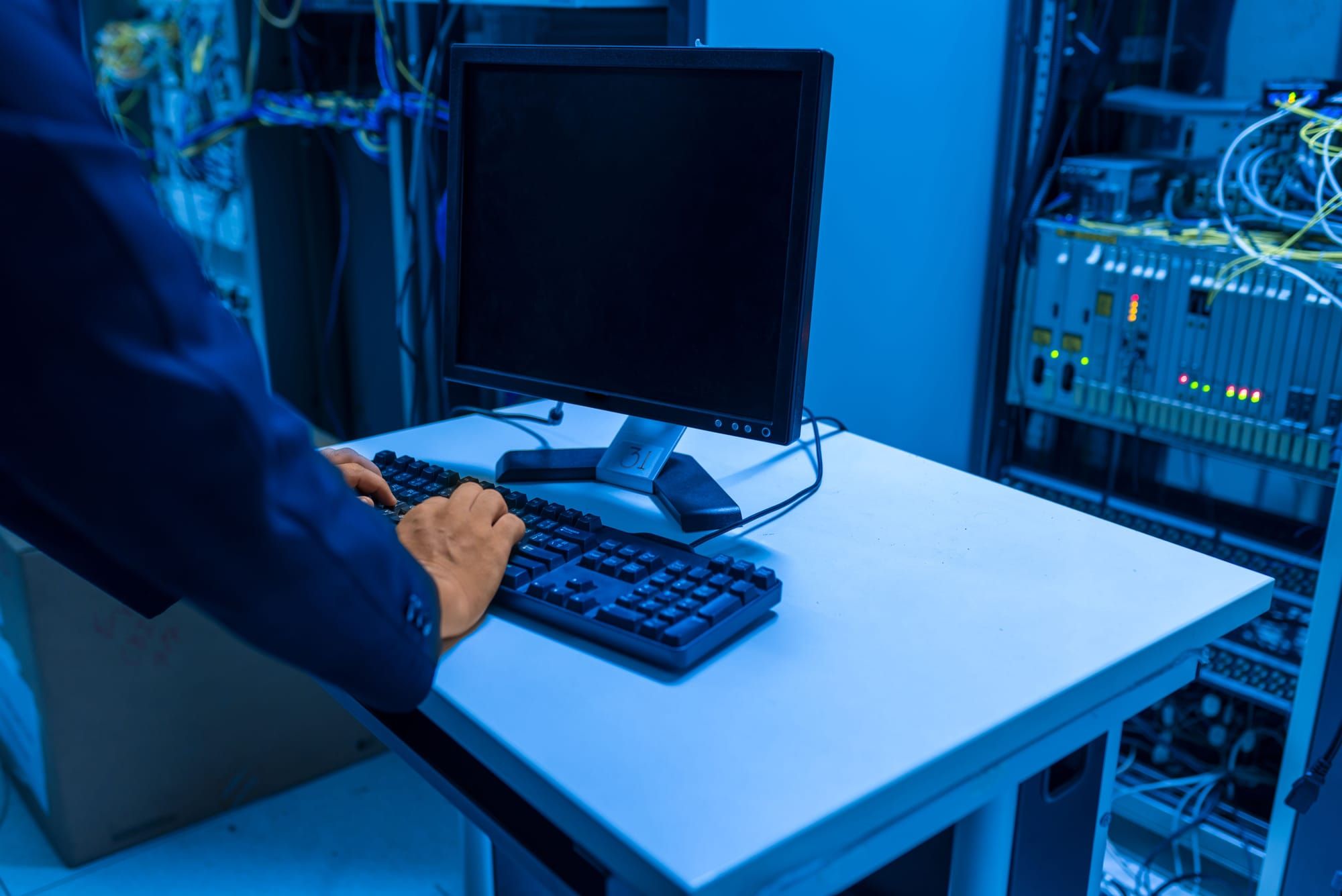 Técnico escribiendo en el teclado de la consola de una sala de servidores, con armarios y cableado al fondo