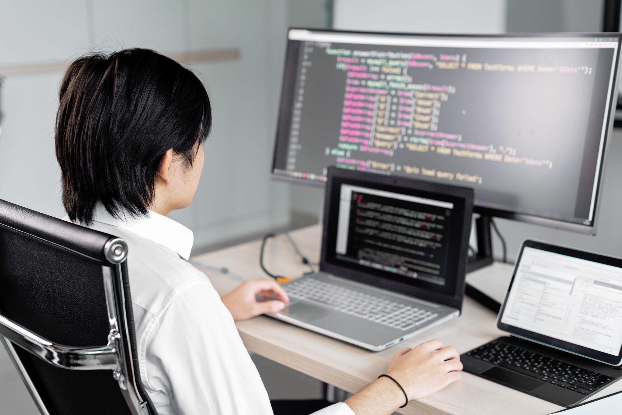 Técnica de espaldas en su puesto, con código en un monitor grande, una terminal en el portátil y una tableta al lado